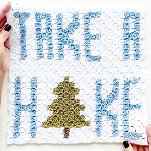 May include: A white crocheted square with the words "Take a Hike" in blue yarn. A green crocheted pine tree is in the center of the square.