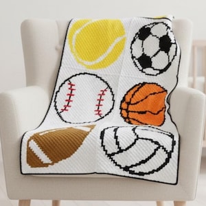 Pode incluir: Um cobertor branco com uma borda preta drapeado sobre uma cadeira. O cobertor apresenta imagens coloridas de bolas esportivas, incluindo uma bola de tênis amarela, uma bola de futebol, uma bola de basquete, uma bola de voleibol, uma bola de beisebol e uma bola de futebol.