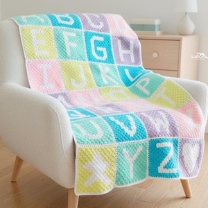 Puede incluir: Una manta de alfabeto de ganchillo en colores pastel sobre un sillón blanco. La manta presenta las letras de la A a la Z en blanco sobre cuadrados rosas, amarillos, azules y morados. En el fondo se ve una cuna de madera.