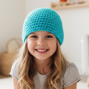 May include: A turquoise crochet beanie hat. The hat is worn by a young person with long blonde hair and a gray shirt. The person is smiling at the camera.