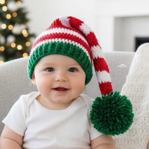 May include: A baby wearing a red, white, and green striped elf hat with a green pom-pom. The hat is crocheted and has a long, pointed tip. The baby is wearing a white onesie and smiling.
