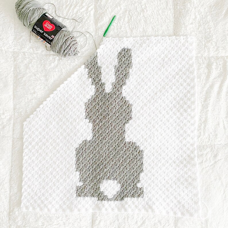 May include: A partially crocheted white blanket featuring a grey rabbit silhouette in the centre. A skein of grey Red Heart Super Saver yarn and a green crochet hook are positioned at the top left corner of the textured, square-stitched fabric.