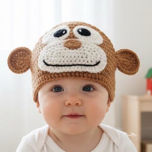 Może przedstawiać: Brązowo-biała czapka w kształcie małpy z dużymi oczami, uszami i uśmiechniętą twarzą. Czapka jest przeznaczona do noszenia na głowie dziecka. Dziecko ma na sobie białe body.