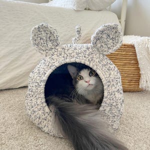 Pode incluir: Uma cama de pelúcia macia para animais de estimação em forma de rato, com grandes orelhas arredondadas e uma pequena cauda encaracolada no topo. A cama é feita de fio salpicado de branco e azul escuro. Um gato de pelo comprido descansa dentro.