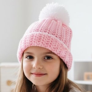 May include: A light pink knitted beanie with a white pom-pom. The hat is worn by a person with long brown hair. The hat is a winter accessory.