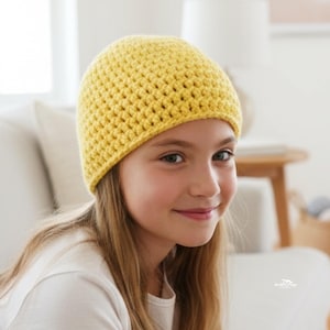 以下が含まれることがあります： 黄色の手編みビーニー帽。帽子は質感のある糸でできており、丸みを帯びた形をしています。長い金髪と白いシャツを着た人が帽子をかぶっています。