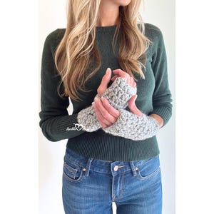 以下が含まれることがあります： グレーの指なし手袋のペア。手袋は、暗い緑色のセーターと青いジーンズを着た人が着用しています。手袋は、テクスチャのあるニットパターンで編まれています。