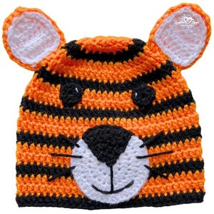 Könnte beinhalten: Ein handgehäkelter Tigerhut in orange und schwarzen Streifen. Der Hut hat ein weißes und schwarzes Tigergesicht mit einem lächelnden Mund und Schnurrhaaren. Die Ohren sind orange mit weißen Innendetails. Ein lustiges und verspieltes Accessoire.