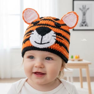 May include: A crochet tiger hat in orange, black, and white, with ear details. The hat features a tiger face design, including a white muzzle and black stripes. The hat is worn by a baby.