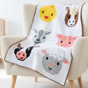 May include: A white crochet blanket featuring colorful farm animal faces: a yellow chick, brown horse, gray goat, pink pig, black and white cow, and gray sheep. The blanket is draped over a cream-colored armchair.