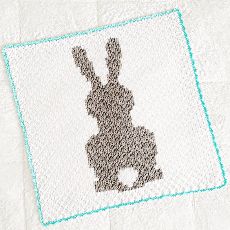 May include: A square crochet blanket featuring a white background with a central silhouette of a rabbit in a textured brown yarn. The blanket is finished with a decorative border in a bright turquoise yarn, creating a distinct contrast.