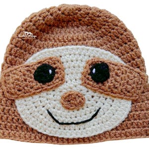 May include: A handmade crochet hat in the shape of a sloth. The hat is primarily brown with a cream-colored face, brown eye patches, and black eyes. The sloth's smiling mouth is outlined in black.
