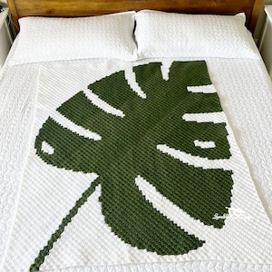 May include: A handmade, white and green crochet blanket featuring a large monstera leaf design. The blanket is displayed on a bed with white bedding and pillows. The blanket's design is a striking contrast of white and dark green.
