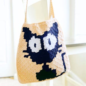 May include: A crocheted tote bag with a black cat design. The cat has white eyes and a black nose.