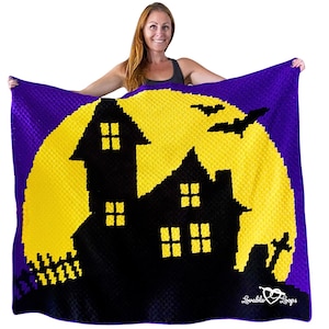 K&ouml;nnte beinhalten: Eine lila, gelbe und schwarze Halloween-Decke mit einer Silhouette eines Spukhauses vor einem gelben Mond. Die Decke hat ein strukturiertes Aussehen und enth&auml;lt zwei schwarze Flederm&auml;use. Die Decke wird von einer Person gehalten.