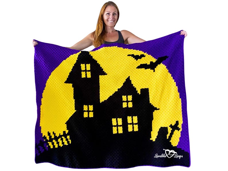 K&ouml;nnte beinhalten: Eine lila, gelbe und schwarze Halloween-Decke mit einer Silhouette eines Spukhauses vor einem gelben Mond. Die Decke hat ein strukturiertes Aussehen und enth&auml;lt zwei schwarze Flederm&auml;use. Die Decke wird von einer Person gehalten.