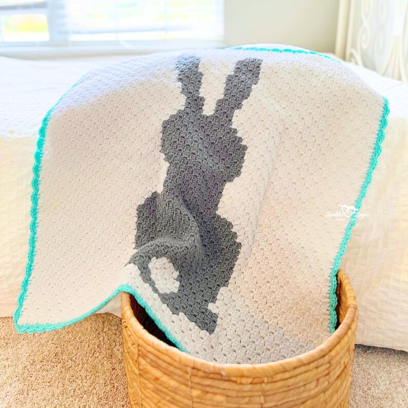 May include: A cream-coloured crochet blanket featuring a large dark grey silhouette of a rabbit in the centre. The blanket has a textured stitch pattern and is finished with a bright turquoise border along the edges. A small logo is visible.