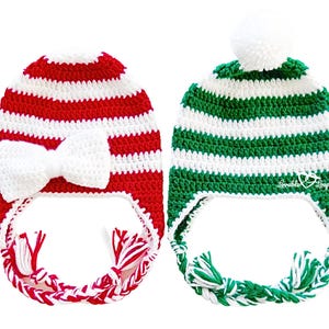 May include: Two handmade crochet hats. One is red and white striped with a white bow and braided ties. The other is green and white striped with a white pom-pom and braided ties. The hats are labeled "Lovable Loops".