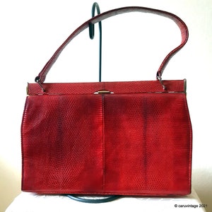 May include: A vintage red handbag with a textured, reptile-like pattern. The bag has a top handle, a rectangular shape, and a gold-coloured clasp. The bag is displayed on a white surface.