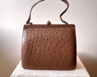 May include: A vintage brown handbag with a textured surface and a top handle. The bag has a rectangular shape with a clasp closure and a small, rounded gold-colored accent. The handle is a darker shade of brown.