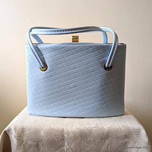May include: A pale blue vintage handbag with a diamond pattern. The handbag has a rectangular shape, a gold clasp, and two handles. The bag is sitting on a cream-coloured tablecloth.