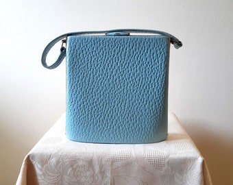 May include: A vintage, sky blue handbag with a textured surface and a matching handle. The rectangular bag has a clasp closure and is displayed on a cream-colored tablecloth.