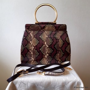 May include: A vintage handbag with a snakeskin pattern in burgundy, brown, and green. It has a gold-tone handle and a detachable shoulder strap. The bag is rectangular with rounded edges.