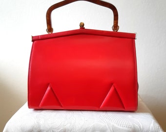 May include: A vintage red handbag with a glossy finish. The bag features a rectangular shape with a tortoiseshell handle and a gold-tone clasp. Two triangular design elements are stitched on the front of the bag.