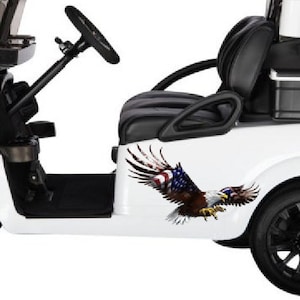 Op de afbeelding: Witte golfkar met zwarte stoelen en een zwart stuurwiel. De kar heeft een zwarte koelbox achterin en een sticker van een adelaar met een Amerikaanse vlag op zijn vleugels.