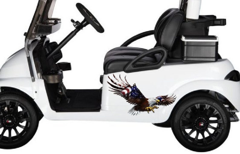 Op de afbeelding: Witte golfkar met zwarte stoelen en een zwart stuurwiel. De kar heeft een zwarte koelbox achterin en een sticker van een adelaar met een Amerikaanse vlag op zijn vleugels.