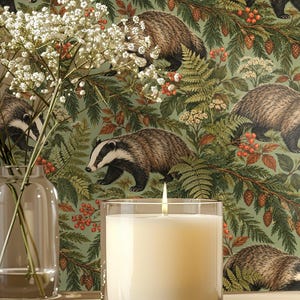 May include: A lit white candle in a clear glass jar sits on a wooden surface in front of a green wallpaper featuring a repeating pattern of badgers, pine branches, ferns, and red berries, alongside a glass vase with white flowers.