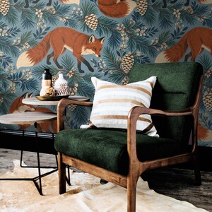 May include: A dark green upholstered armchair with a wooden frame and a white and tan striped throw pillow. Beside the chair is a small round side table holding a black bottle and a white patterned vase against fox-patterned wallpaper.