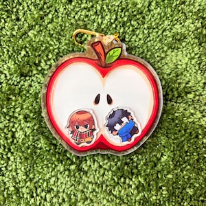 Peut inclure: Un porte-clés transparent en forme de pomme avec un contour rouge et deux personnages de dessins animés à l'intérieur. Le porte-clés a un fermoir doré et est posé sur un fond vert texturé.
