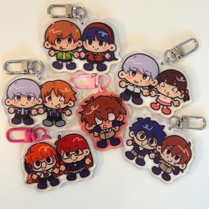 Op de afbeelding: Set van 8 kleurrijke cartoon-personage sleutelhangers. Elke sleutelhanger heeft twee personages die elkaar vasthouden. De personages zijn getekend in een chibi-stijl en hebben verschillende haarkleuren, kleding en uitdrukkingen.