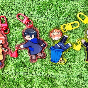 May include: Four acrylic keyrings featuring anime-style characters with red and yellow clasps. The characters are in various poses, including one in a bin. The background is a green, textured surface.