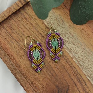 May include: A pair of handmade earrings featuring a green stone and purple, green, and gold macrame. The earrings are displayed on a wooden surface.