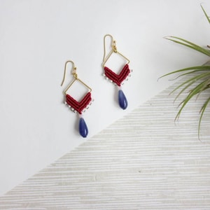 May include: Red and white string earrings with a blue teardrop-shaped stone hanging from each earring. The earrings are made with a gold-coloured wire frame.