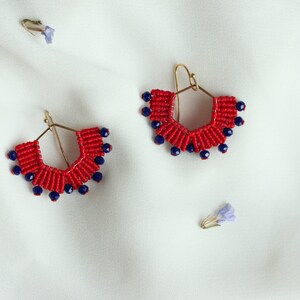 May include: A pair of red and blue beaded earrings with gold hoops. The earrings are in the shape of a crescent moon and are hanging against a white background.