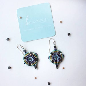 May include: A pair of turquoise, purple, gold and black macrame earrings with black beads and silver hooks. The earrings are handmade and feature a geometric design. The earrings are displayed on a white surface with a business card that reads "knotorious jewellery - handcrafted macrame & more..."