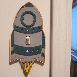 May include: A light switch cover shaped like a rocket ship with a gray body, teal accents, and a yellow flame. The switch is white.