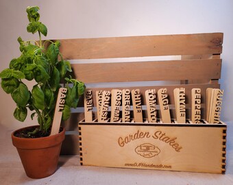 May include: A set of wooden garden stakes with labels for different herbs and vegetables, including basil, squash, rosemary, oregano, chive, tomato, sage, thyme, peppers, and zucchini. The stakes are organized in a wooden box with the text "Garden Stakes" and a website address.