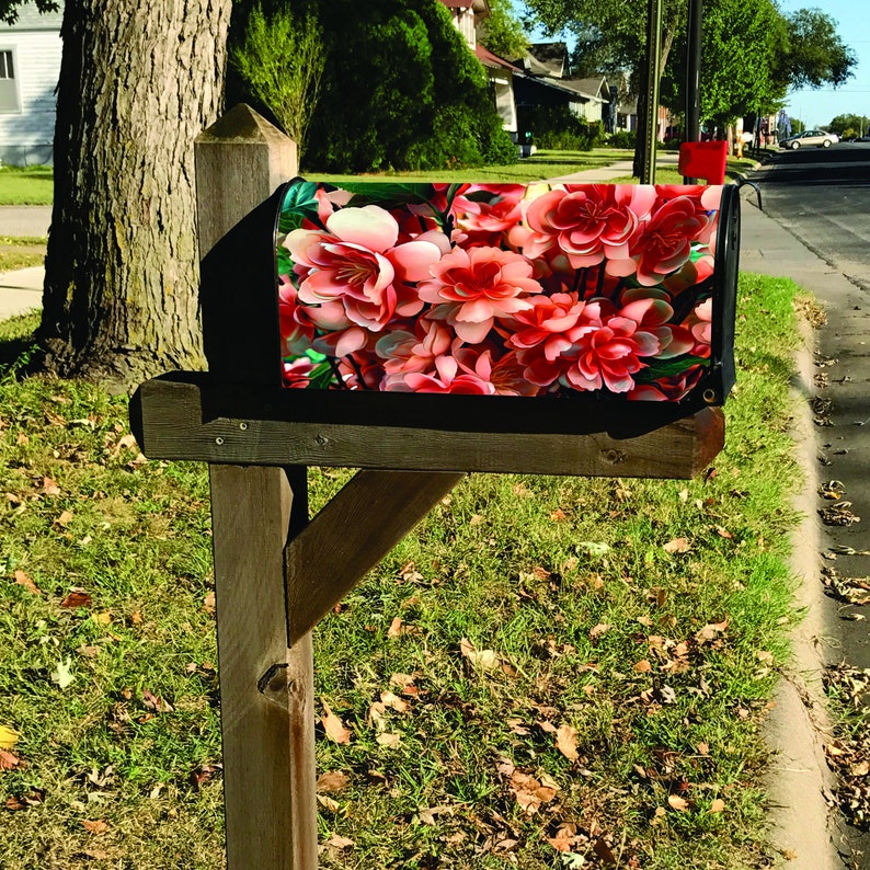 May include: A wooden mailbox with a pink floral design. The mailbox is mounted on a wooden post and is surrounded by green grass and fallen leaves.