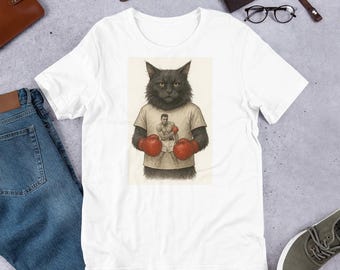 Puede incluir: Camiseta blanca con un gato negro que lleva una camiseta de boxeador y guantes rojos. El gato tiene una expresión seria. La camiseta está sobre una superficie gris claro con otros accesorios.