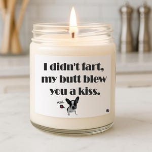May include: A lit, white candle in a glass jar. The label reads "I didn't fart, my butt blew you a kiss." A cartoon dog with a red rose is on the label. The candle is on a white surface.