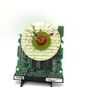 May include: A clock made from a repurposed computer hard drive circuit board. The clock face is a white disc with green and red accents. The clock hands are gold and red.
