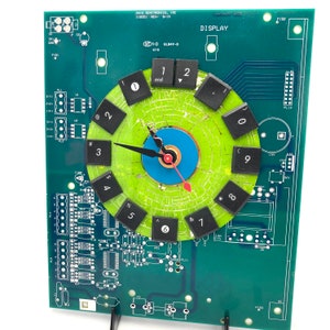 May include: A unique clock made from a green circuit board. The clock face is a circle with black keycaps as numbers. The hands are black and red. The clock is mounted on a black stand.