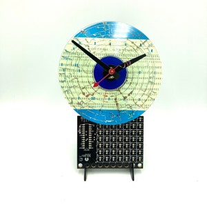 May include: A clock with a blue and white cracked vinyl record face. The clock face has the text "FORTRAN STATEMENT" and numbers around the edge. The clock is mounted on a black circuit board with a stand.