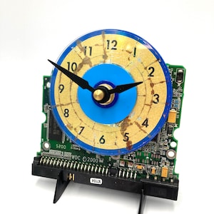 May include: A clock made from a repurposed computer hard drive. The clock face is a blue and gold circle with black numbers and hands. The hard drive is green and has the text "WDC © 2000" and "V7154" printed on it.