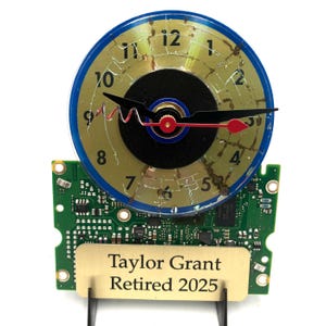 May include: A gold and blue clock with a cracked CD face, mounted on a green circuit board. The clock has a black hand and a red second hand. A gold plaque below reads "Taylor Grant Retired 2025".