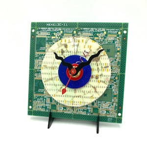 May include: A clock made from a circuit board with a white face and black hands. The clock has a blue and red ring around the centre. The circuit board is green and has the text "H64613C-I11" and "WALCHEM" on it.